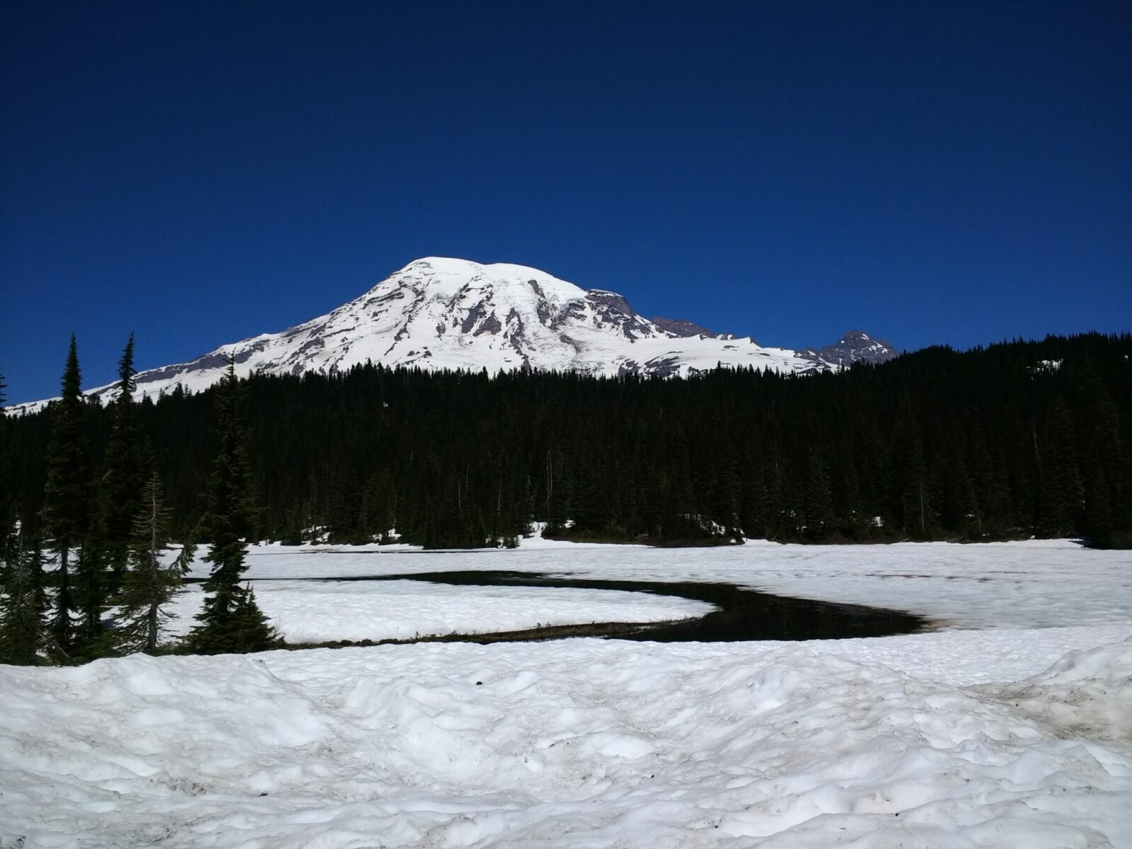 A large snow covered mountain behind forested hills. A partially frozen lake in the foreground