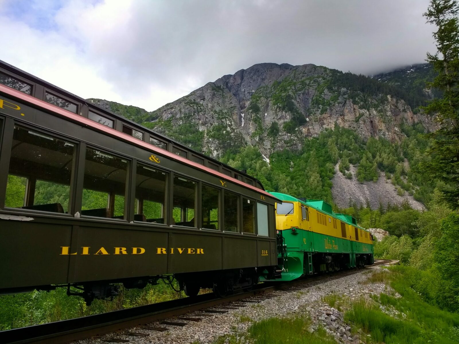 A green and yellow diesel train engine pulling a historic brown train car with a red roof. The train track is going through the forest and by mountains