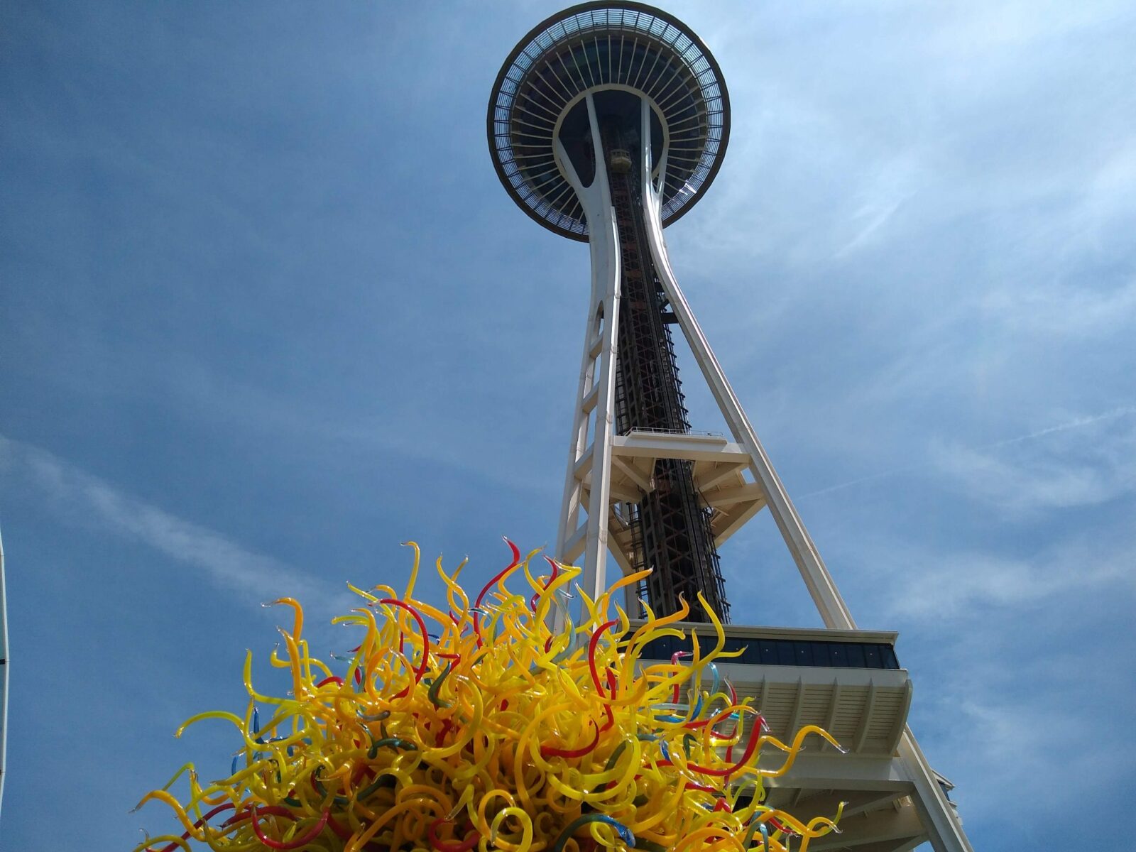 A bright yellow, orange and red glass sculpture at the base of the Space Needle on a sunny day looking up at the Space Needle from below