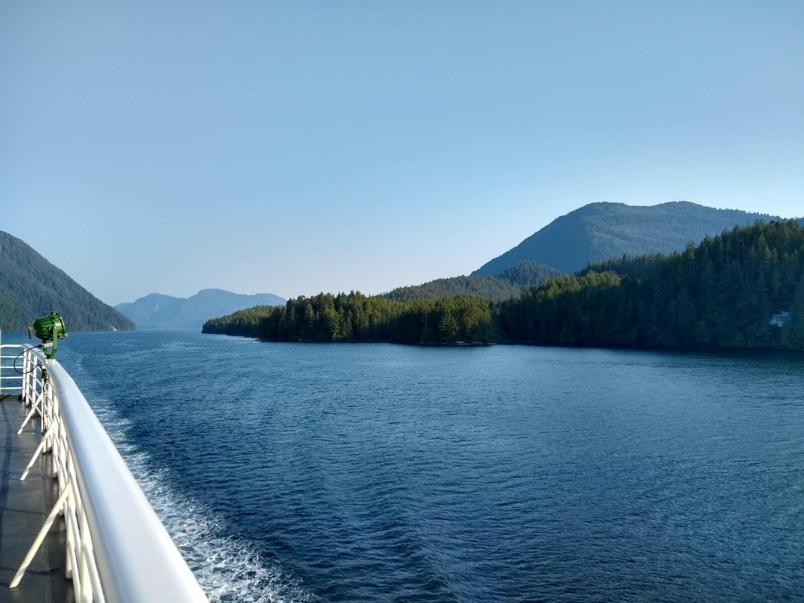 The deck of a ship in a narrow passage of water between forested hillsides and islands.