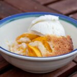 A white bowl with a blue rim contains a piece of peach cobbler and a scoop of vanilla ice cream