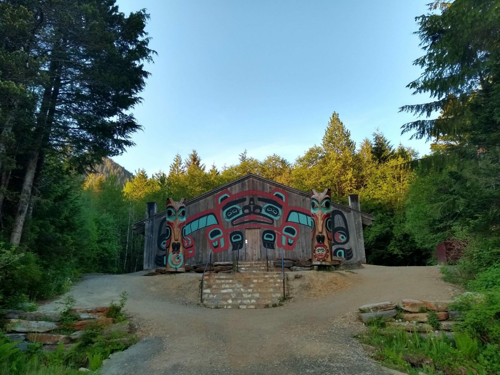 The house screen on the outside of a native clan house in Saxman near Ketchikan. the building is surrounded by forest and the sun is setting.