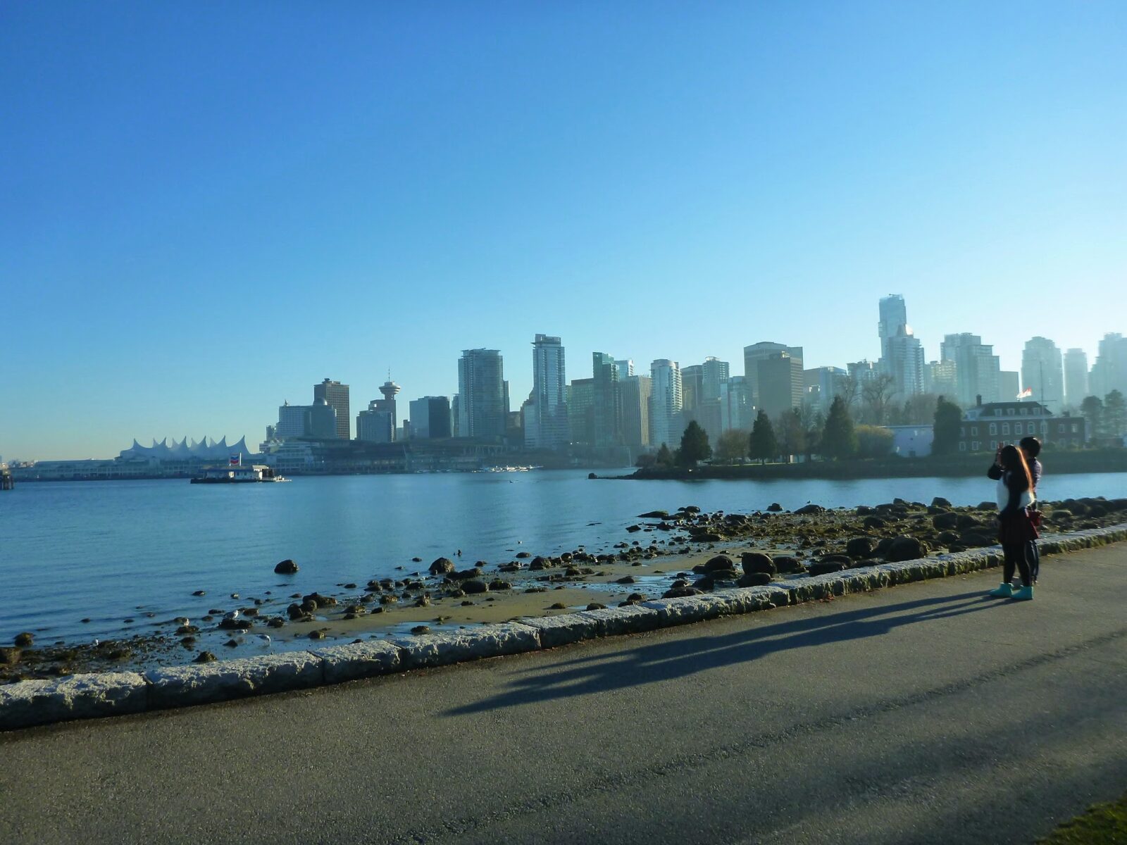 A city skyline is seen in the distance across a body of water. In the foreground is a paved trail and a rocky beach. It's a blue sky sunny day