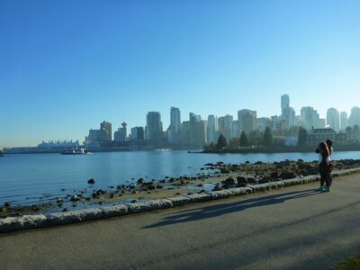 A city skyline is seen in the distance across a body of water. In the foreground is a paved trail and a rocky beach. It's a blue sky sunny day
