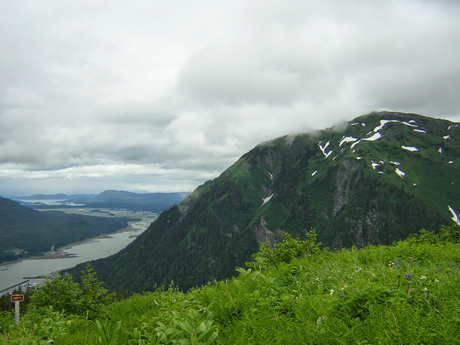 A meadow in the foreground and green mountains around on a ridge. In the distance there is water far below and more mountains