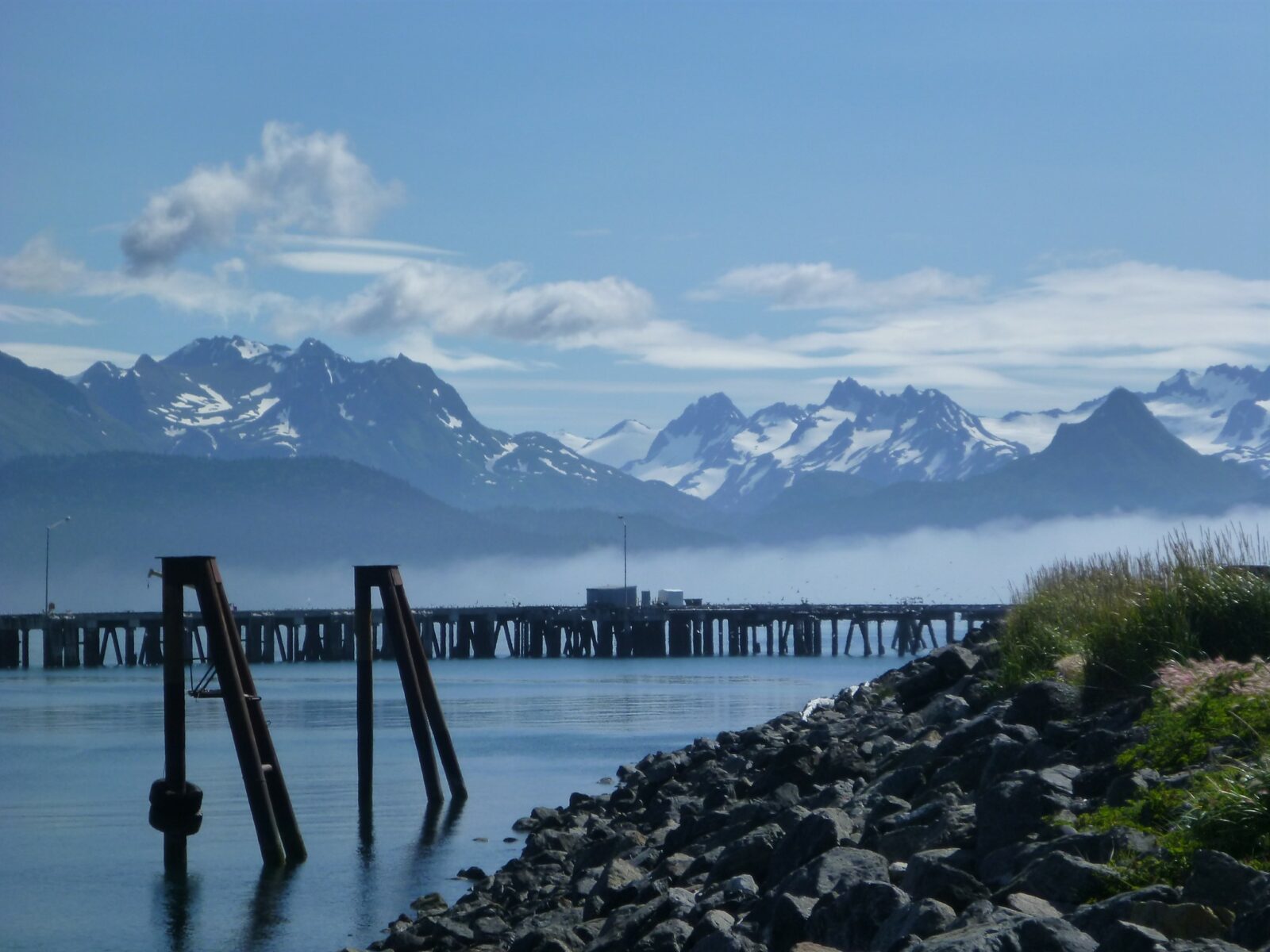 Low fog surrounds a dock and a rocky shore on an otherwise sunny day on the Kenai Peninsula. In the distance are big mountains with lingering snow and glaciers