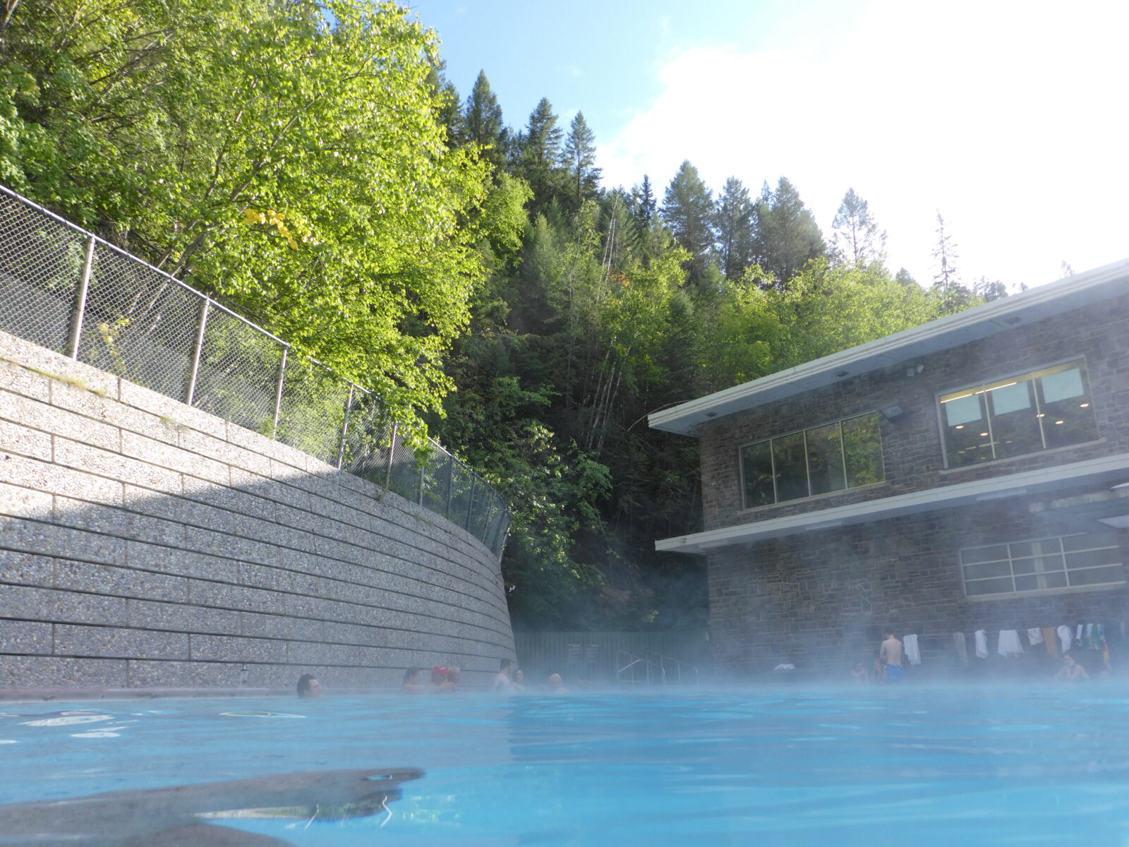 A hot springs pool with a few people in it. There is a stone wall around the pool and a forest above. There is also a building next to it