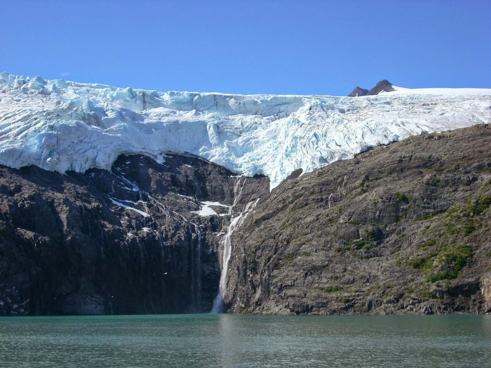 A hanging glacier over rocks next to the water with a small waterfall coming down on a glacier cruise, one of the best day trip tours in Alaska