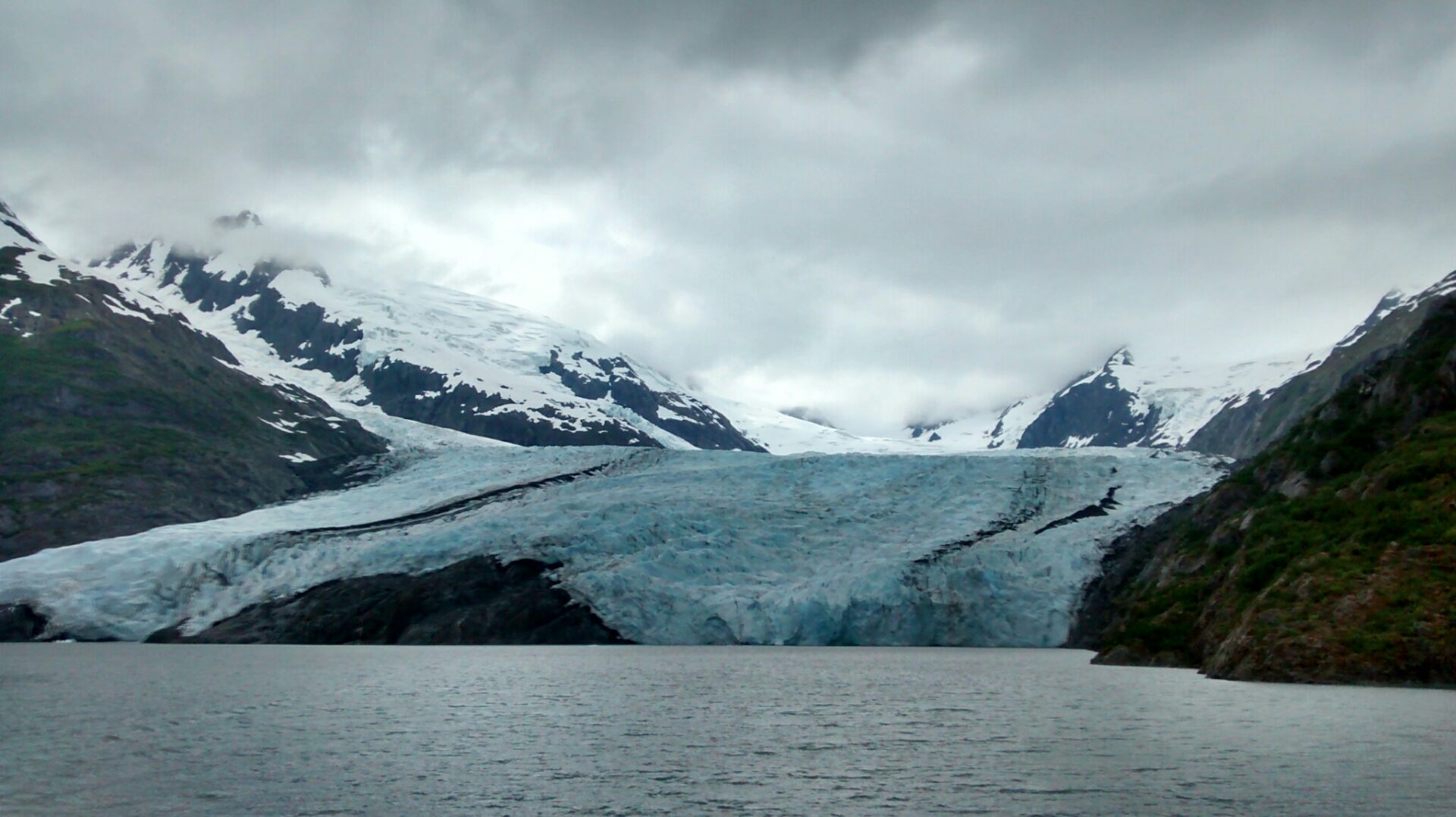 A glacier coming down to touch a lake between rocky hills and snowy mountains on a cloudy day