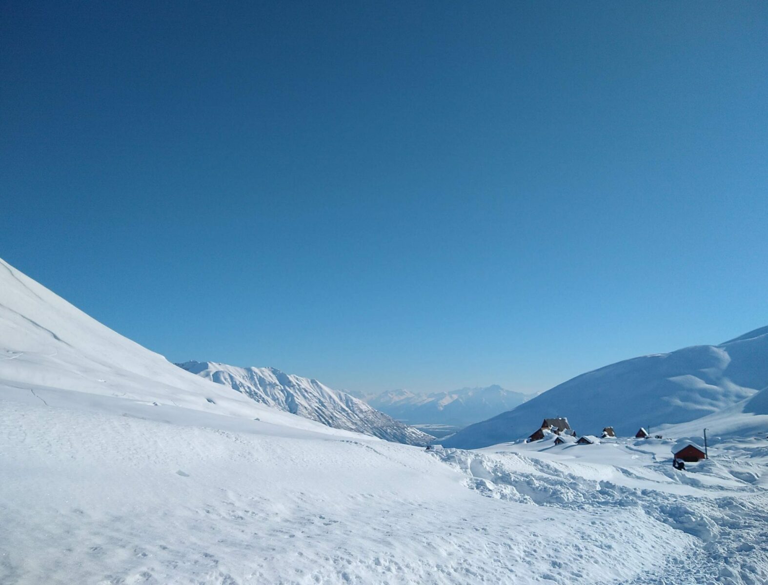 A snowy pass with a few buildings almost covered in snow on a sunny day with a view to distance water and even more mountains