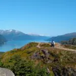 A hiker stands on a ridge above a fjord and distant mountains on a sunny day