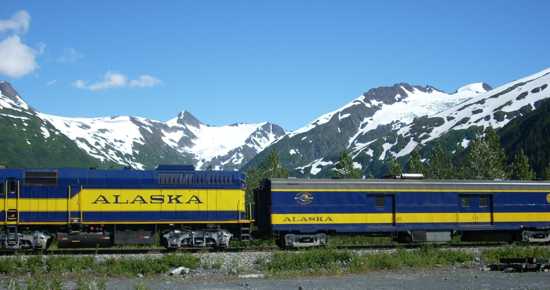 An Alaska railroad engine and baggage car. The cars are blue with yellow stripes that say Alaska. There are glaciers and mountains behind the train.