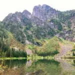A small glassy alpine lake with trees and mountains reflected in it. The lake is surrounded by high mountains and evergreen trees and rocks