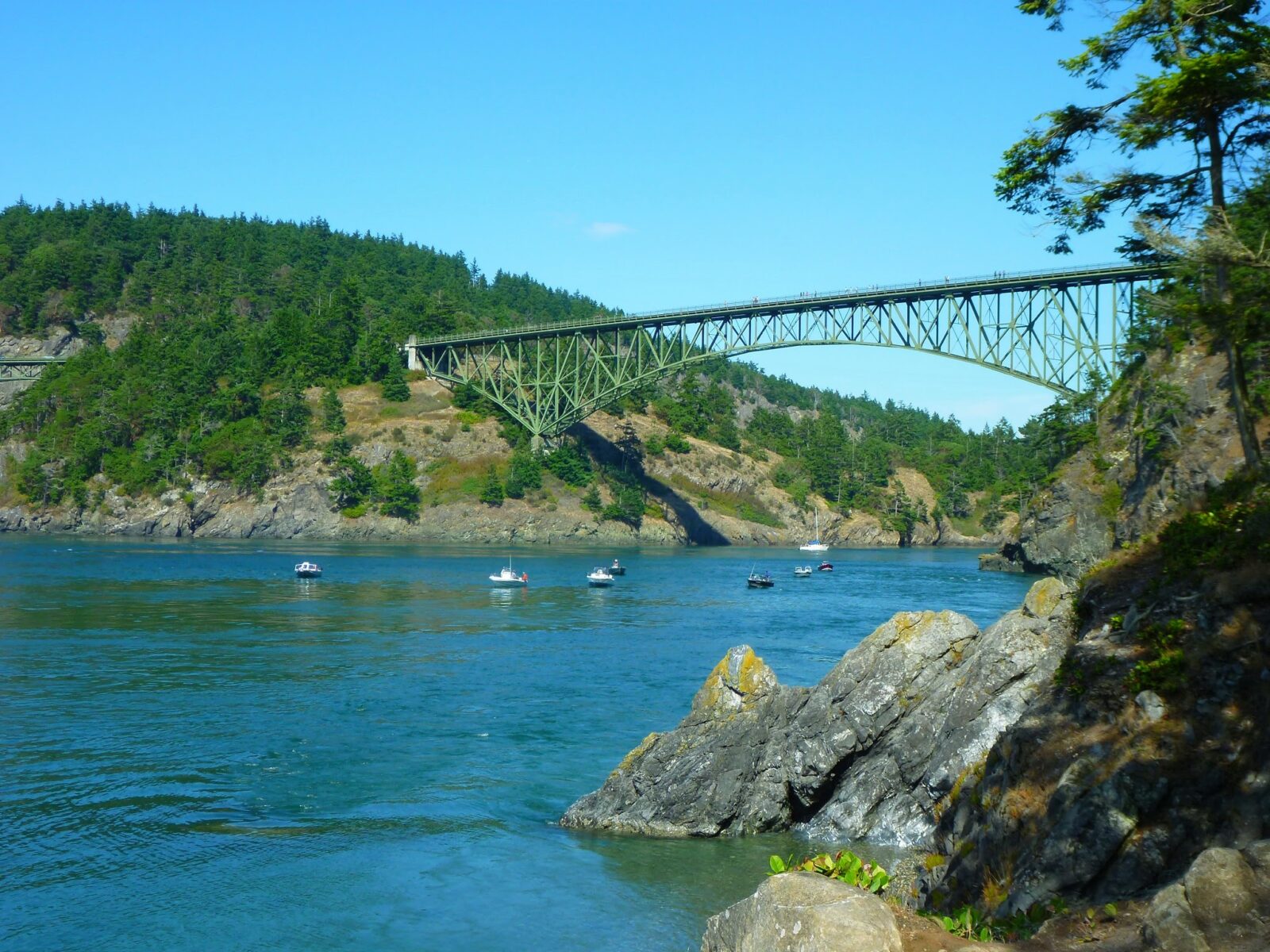 A green metal historic trestle bridge over the water between rocky and forested shore. There are several fishing boats and a sailboat under the bridge