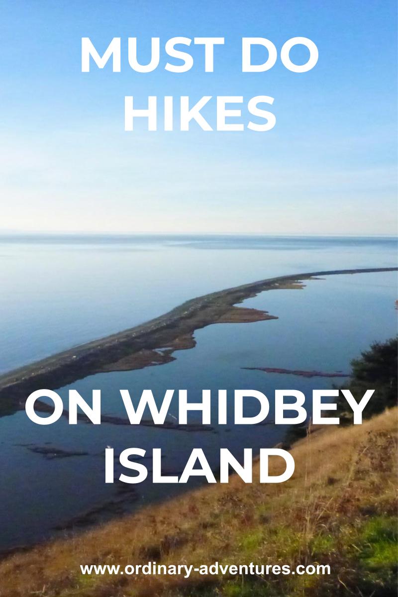 A sand spit on a beach extends away from a grassy hillside encircling a lagoon. Beyond there is water in the distance on a sunny day. Text reads: must do hikes on whidbey island