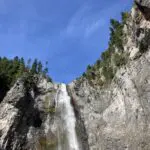 looking up at a high waterfall coming over a cliff with evergreen trees on both sides of the cliff on a sunny day
