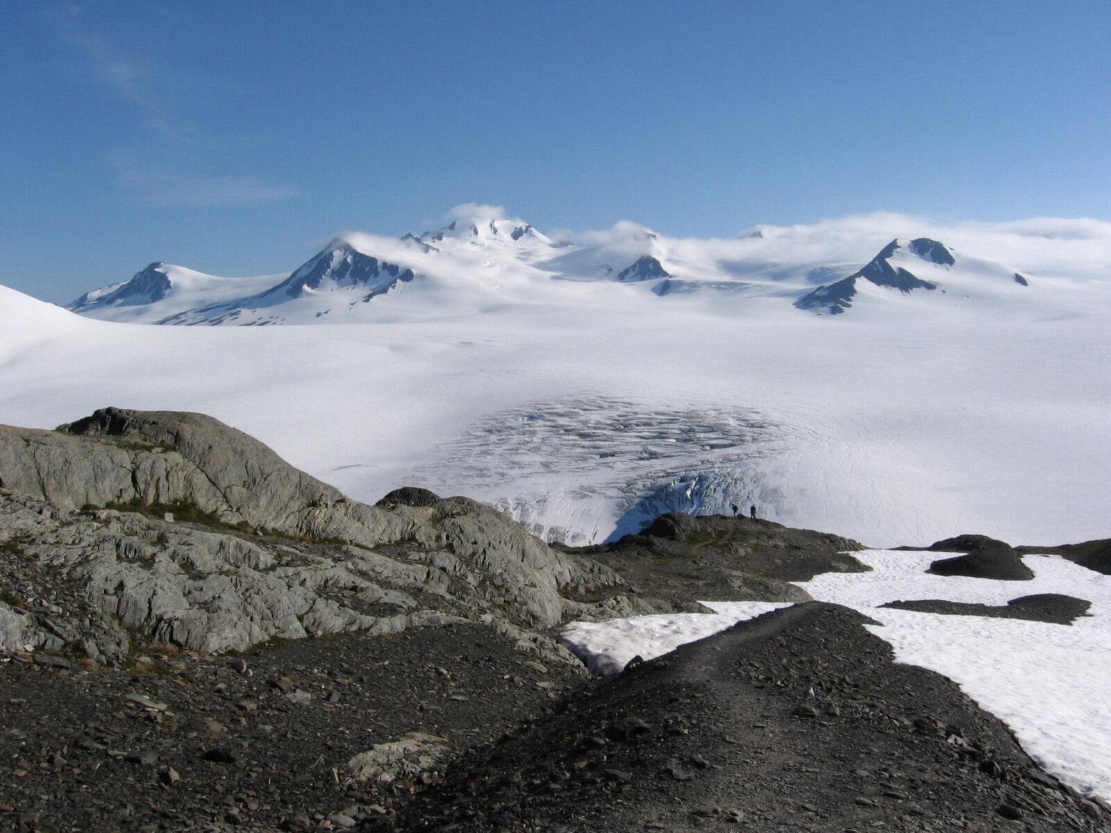 Two hikers in the distance near rocks and a snow field at the end of the Harding icefield hike. A glacier in the distance and mountains partially covered in clouds.