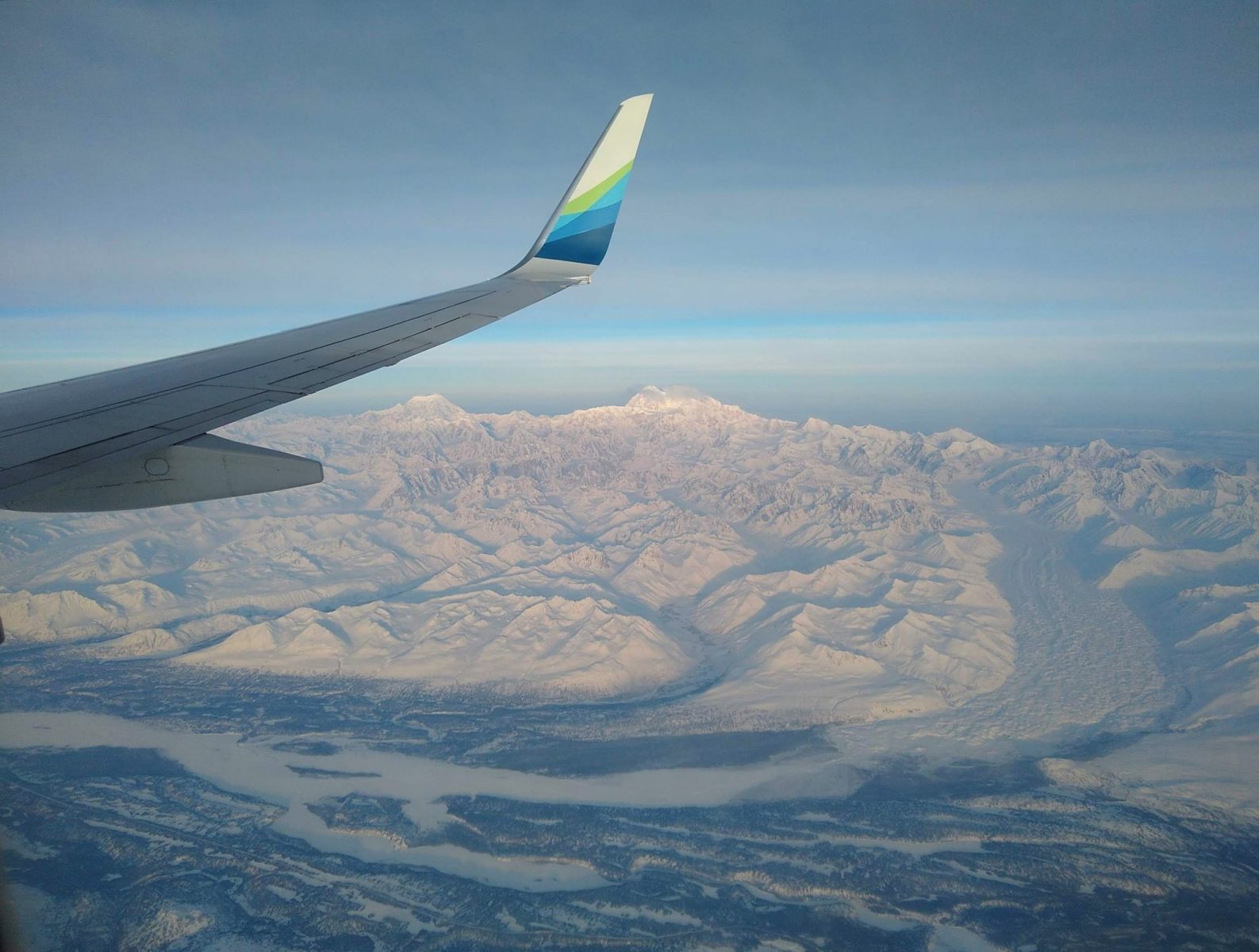 an airplane wing over high mountains and glaciers and a river below