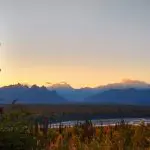 Sunset behind Denali and other mountains of the Alaska range, camping in Alaska at Denali State park. The Susitna River flows through the middle of the image and there are evergreen and fall colored shrubs in the foreground.