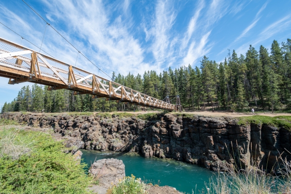  Un pont piétonnier en bois et métal traversant une rivière turquoise dans un canyon rocheux. Il y a des arbres à côté du canyon 