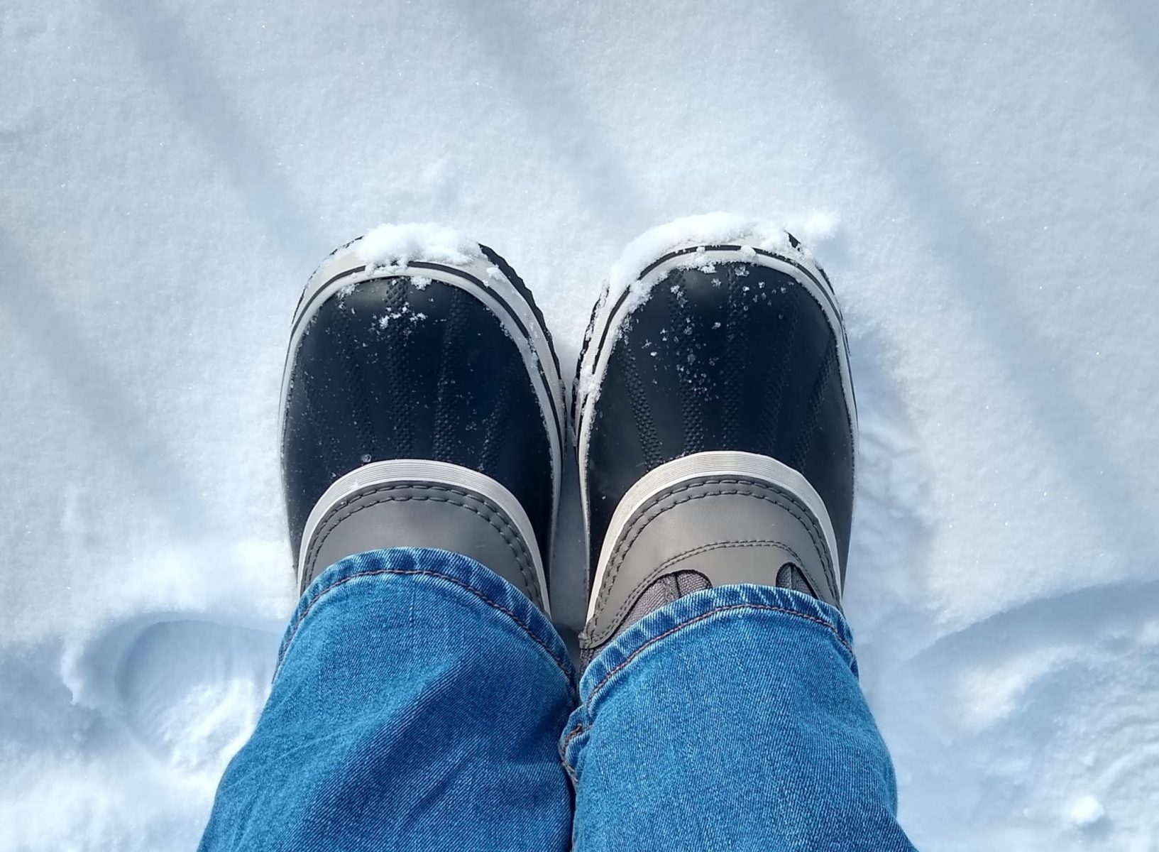 Best boots for Alaska in winter, the bottoms of winter snowboots are visible under jeans seen from above. The boots have a navy blue sole and a gray middle section. The person wearing them is standing in the snow