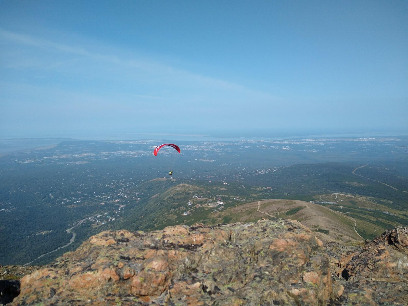 The rocky summit of flattop mountain with the city of Anchorage far below in the distance. A trail and some roads, houses and forests are also seen far below. In the foreground is a paraglider with a red parachute