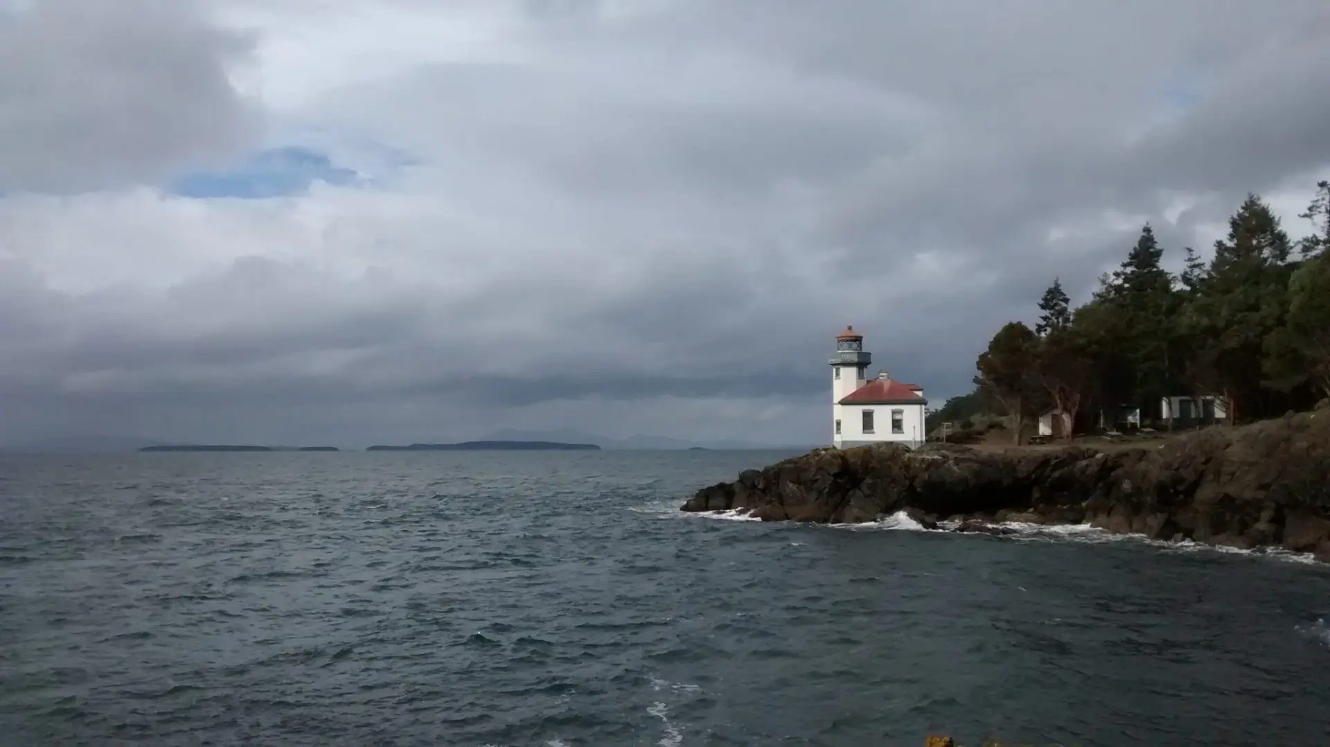 A white and red lighthouse on a rocky shore of San Juan Island next to the forest on an overcast day