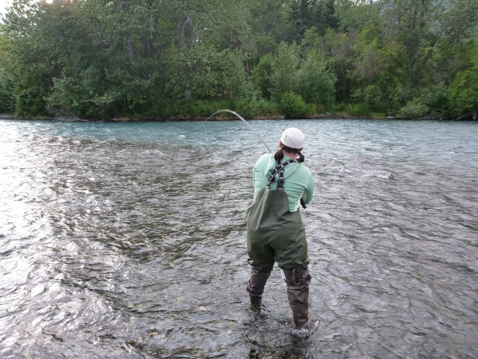 A woman fishing in a river with a hat, waterproof green overalls and brown hip waders