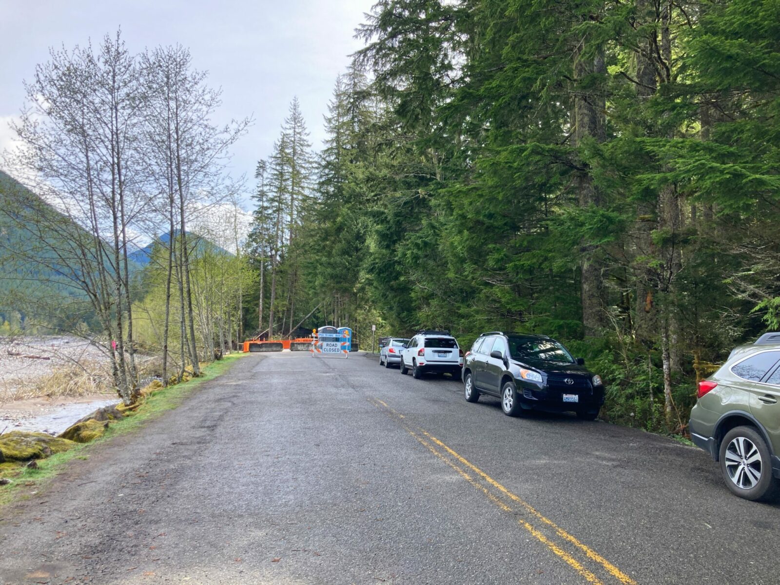 A two lane paved road ends at a barricade with a road closed sign and two port potties. Four cars are parked along the side of the road. There is a forest, hillsides and a river along the side of the road.