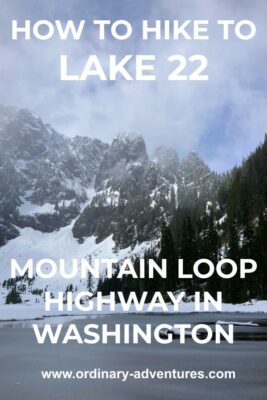 Snowy rocky mountains around a partially frozen lake. There is also forest around one side of the lake, and there are clouds and sun around the mountains. A couple of logs with snow on them are sticking out into the lake. Text reads: How to hike to Lake 22 Mountain loop highway in Washington