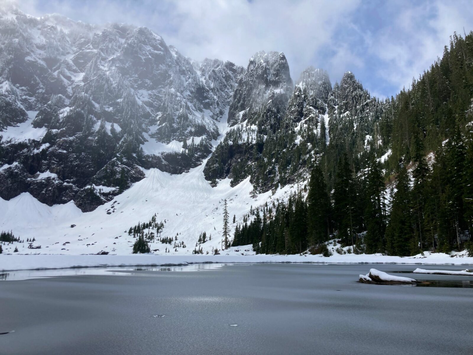 Snowy rocky mountains around partially frozen Lake 22. There is also forest around one side of the lake, and there are clouds and sun around the mountains. A couple of logs with snow on them are sticking out into the lake