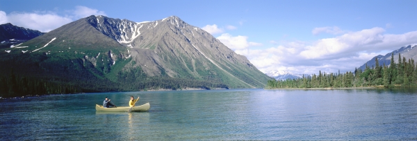  Deux personnes pagayent en canoë sur un lac par une journée principalement ensoleillée. Le lac est entouré de forêts et de montagnes 