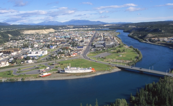 An aerial view of the city of Whitehorse in the Yukon Territory. The blue Yukon River is on the side of town and a bridge crosses it near a historic riverboat. There are hills and mountains in the background