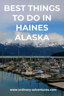 small boat harbor with many boats docked inside a breakwater. Across the water are high mountains with snow and some clouds around them on a partly sunny day. Text reads: best things to do in Haines Alaska