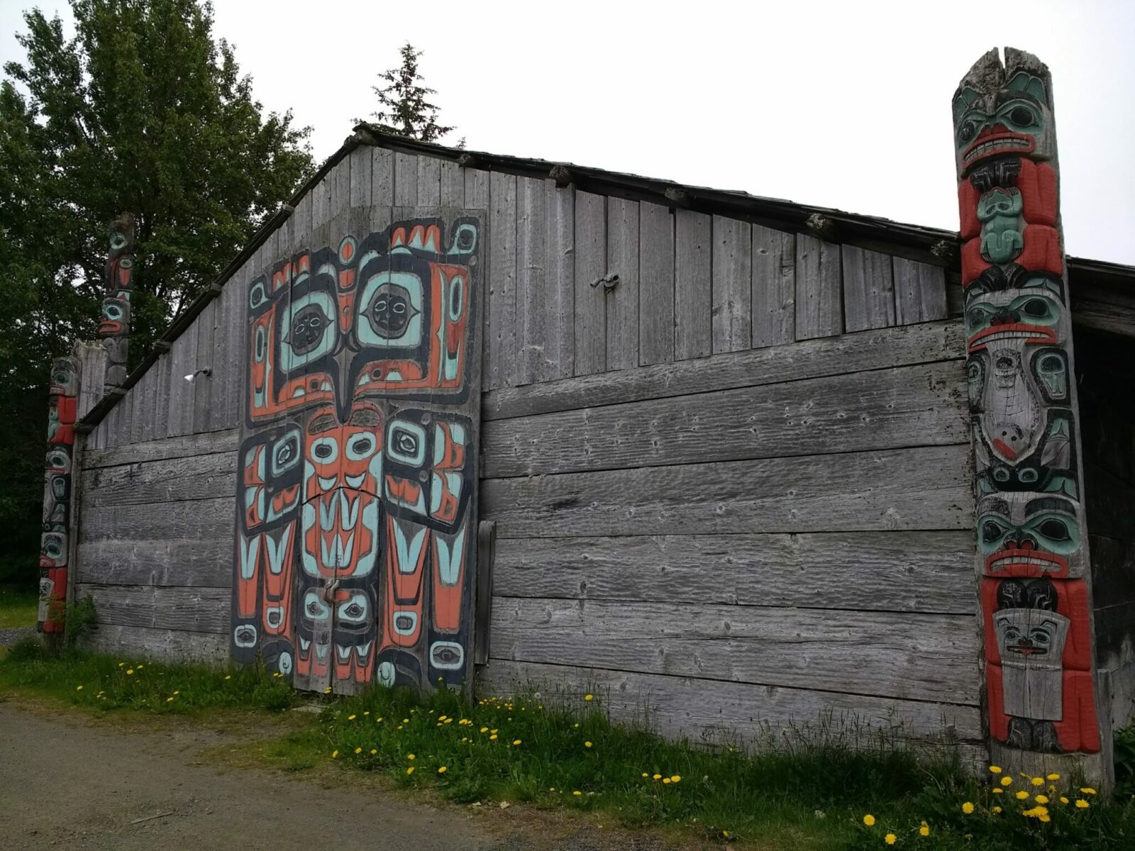 A traditional Chilkat tribal house. It is large, one story and made of weathered wood. On the outside is a house screen, a large flat piece of art that goes on a wall. Next to it are two house poles, which are like totem poles but shorter and on the corners of a house. There is also a totem pole behind the house next to a tree