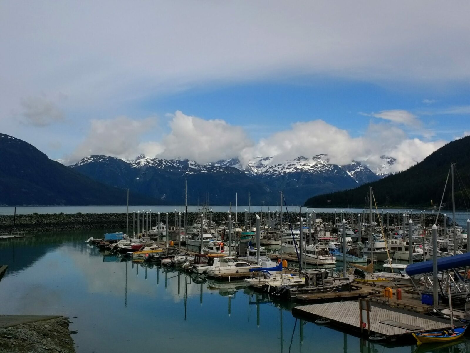 The Haines Alaska small boat harbor with many boats docked inside a breakwater. Across the water are high mountains with snow and some clouds around them on a partly sunny day