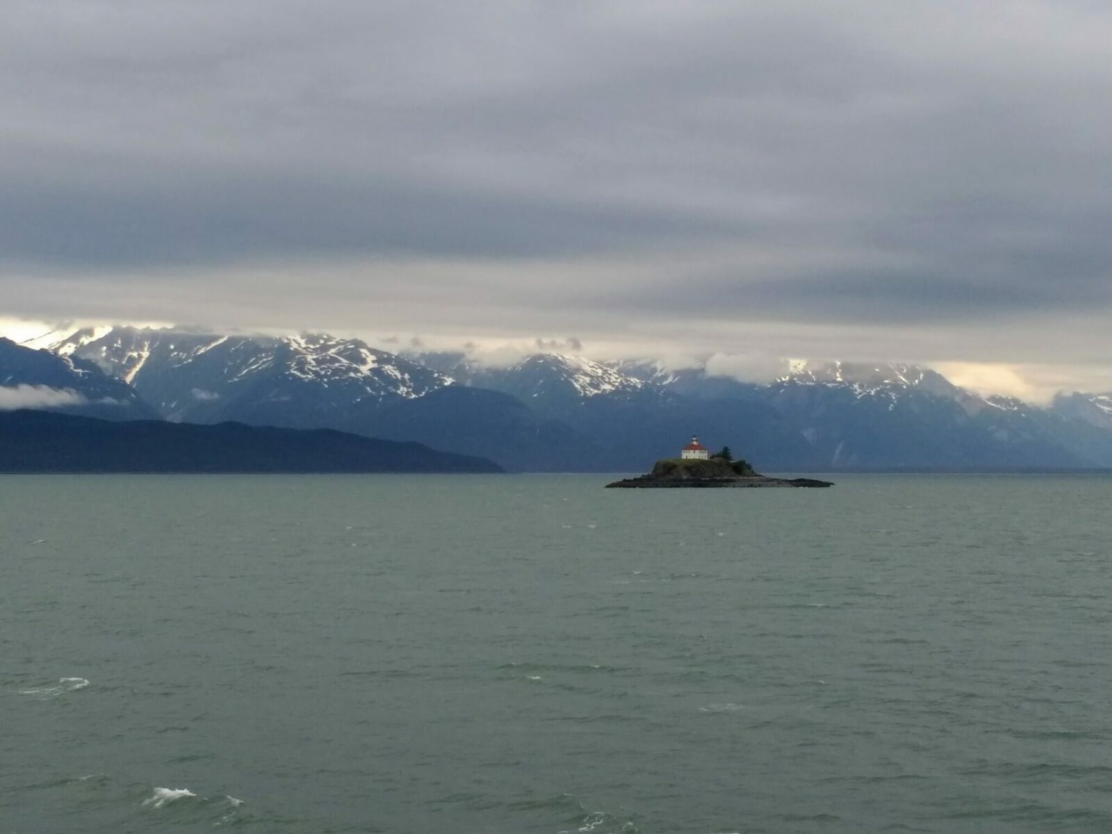 A lighthouse in a white building with a red roof on a small rocky island in the Lynn Canal in Alaska. Around the island is water and forested hills and snow capped mountains on a cloudy day