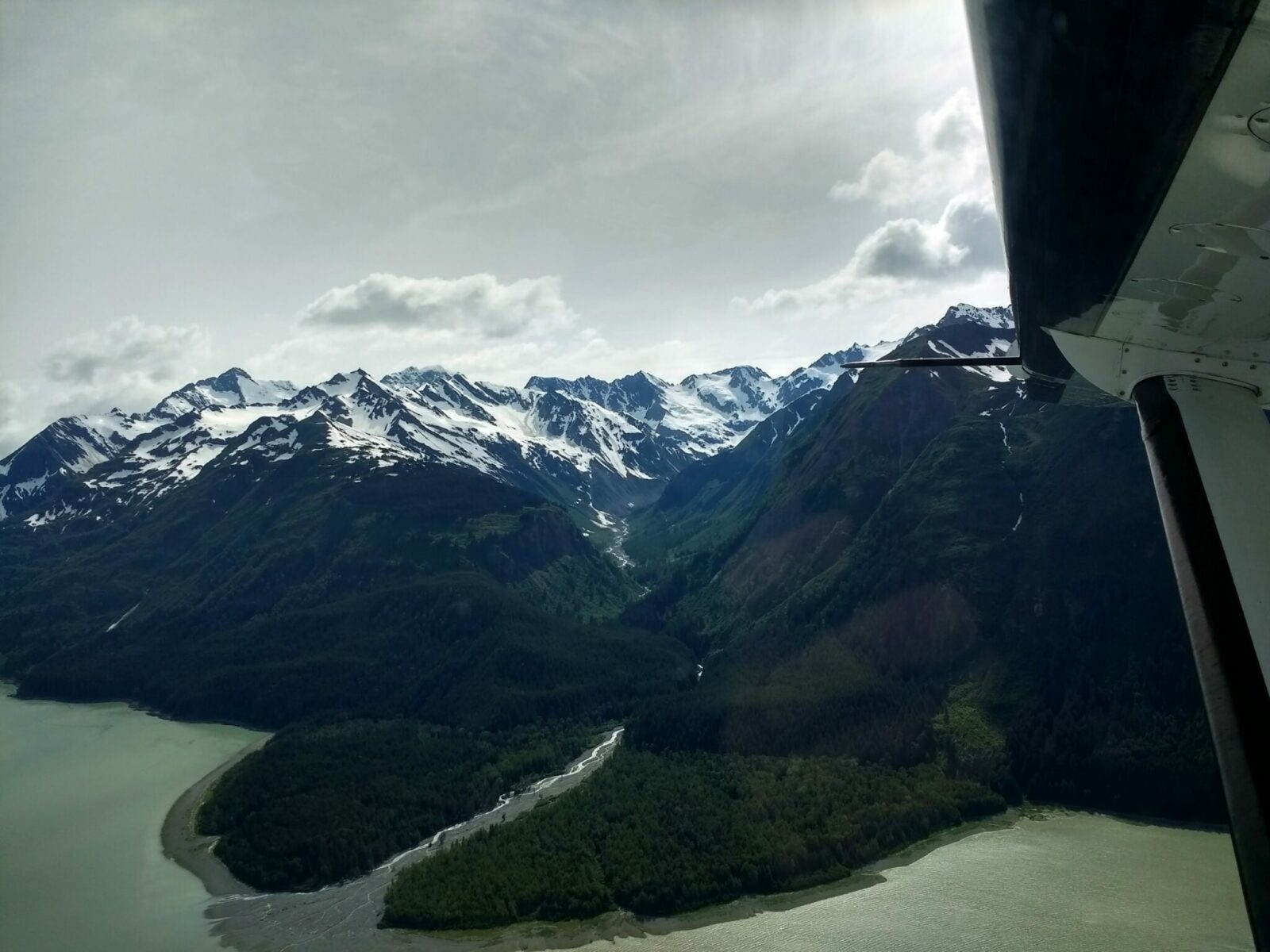 An airplane wing seen from inside a small plane between Haines and Juneau Alaska. Out the window of the plan there is a river coming into the ocean through a forested valley. High snowy mountains and glaciers are above the valley on a partly cloudy day