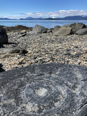 An ancient petroglyph on a dark rock in the foreground. It is a circle with circles around it. It's on a gravel beach with other larger rocks. In the distance are mountains and a few clouds