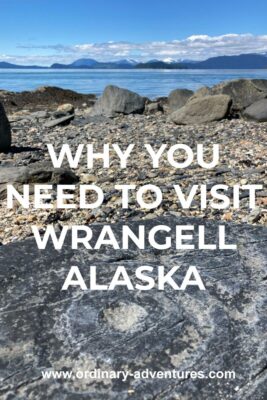 An ancient petroglyph on a dark rock in the foreground. It is a circle with circles around it. It's on a gravel beach with other larger rocks. In the distance are mountains and a few clouds. Text reads: Why you need to visit Wrangell Alaska