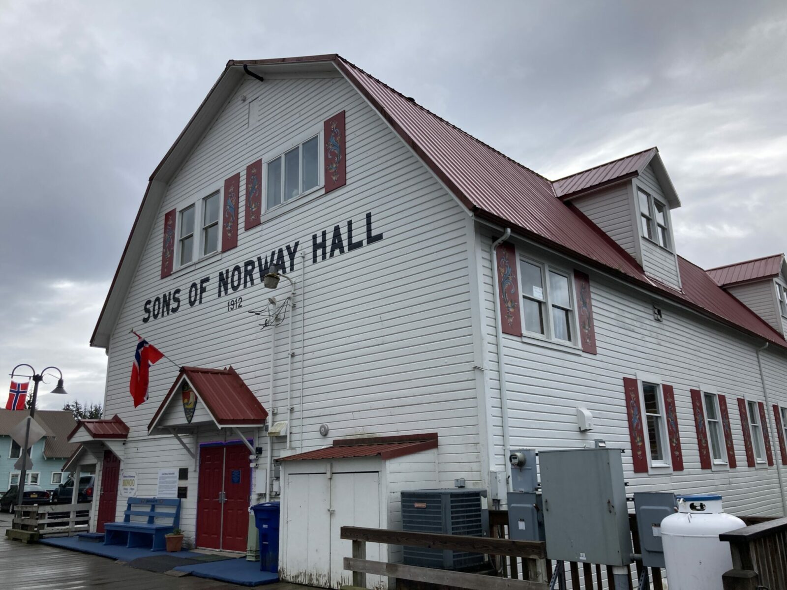 A large barn shaped white wooden building with a red roof. The building says Sons of Norway Hall and has two Norwegian flags