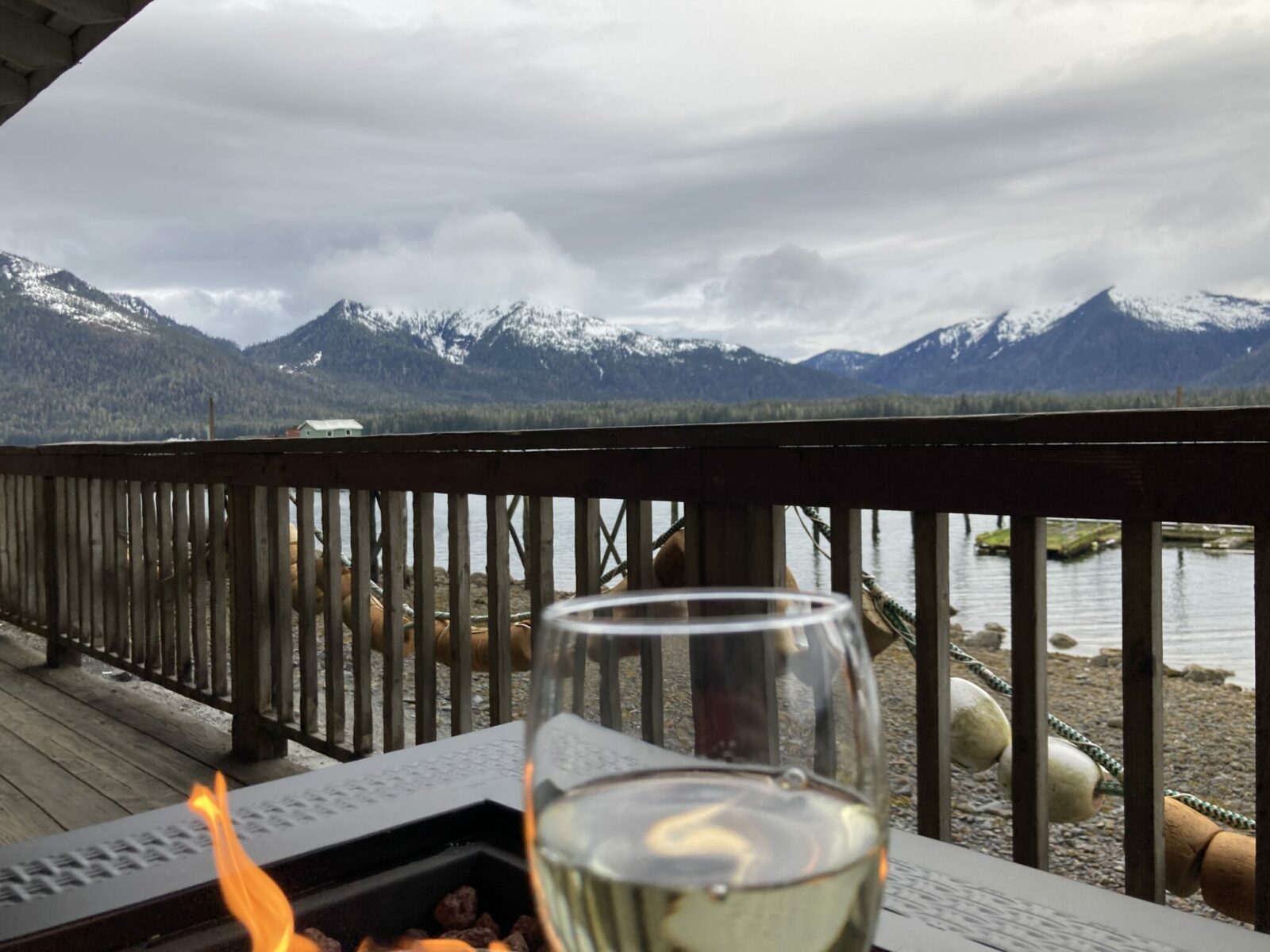 distant mountains with snow and forested lowlands. In the foreground there is a propane firepit on a deck with buoys and a beach at low tide. An out of focus wineglass is in the front