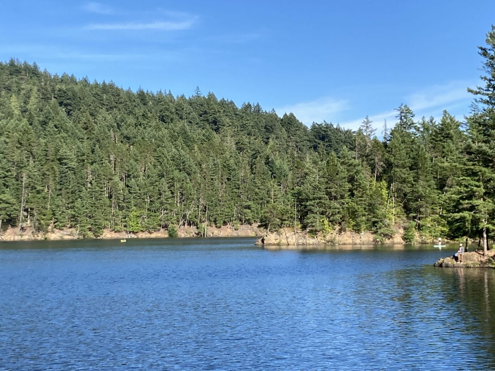 A part of a blue lake surrounded by rocks and evergreen forest on a sunny day. There is one yellow canoe in the water and a stand up paddleboarder. One person is sitting on the shore