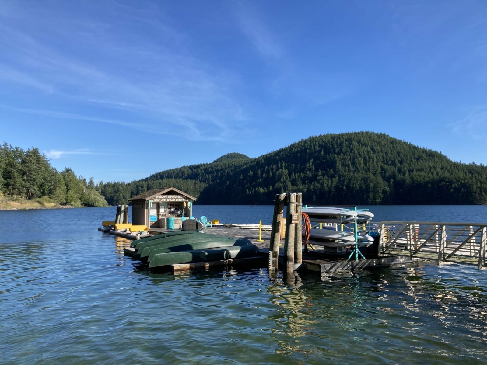 A blue lake surrounded by forest on a sunny day. In the foreground is a dock with several green canoes, a rack of paddleboards and three blue deck chairs. There is a small wooden structure advertising boat rentals