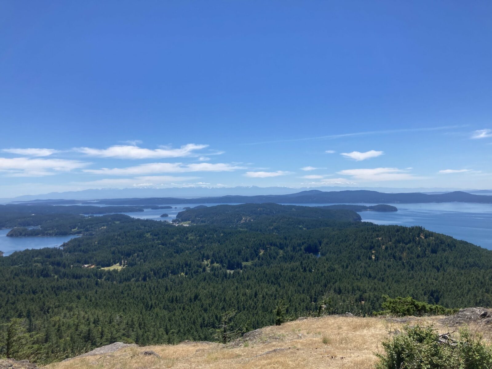 One of the best Orcas Island hikes, Turtlehead summit is a grassy and rocky small meadow that looks out over the Salish Sea in Washington State. There are forested islands and water below, stretching all the way to distant snow capped mountains on a mostly sunny day with a few clouds.