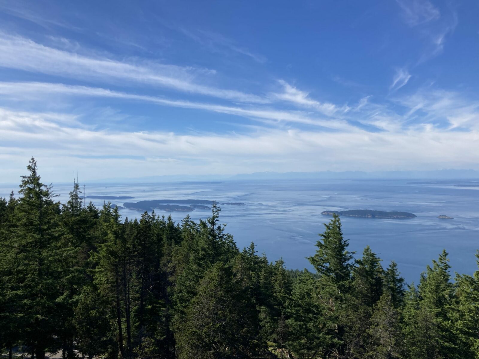 Several islands in the water below the summit of Mt Constitution on Orcas Island. There is a forest hillside in the foreground and there are some thin clouds. 