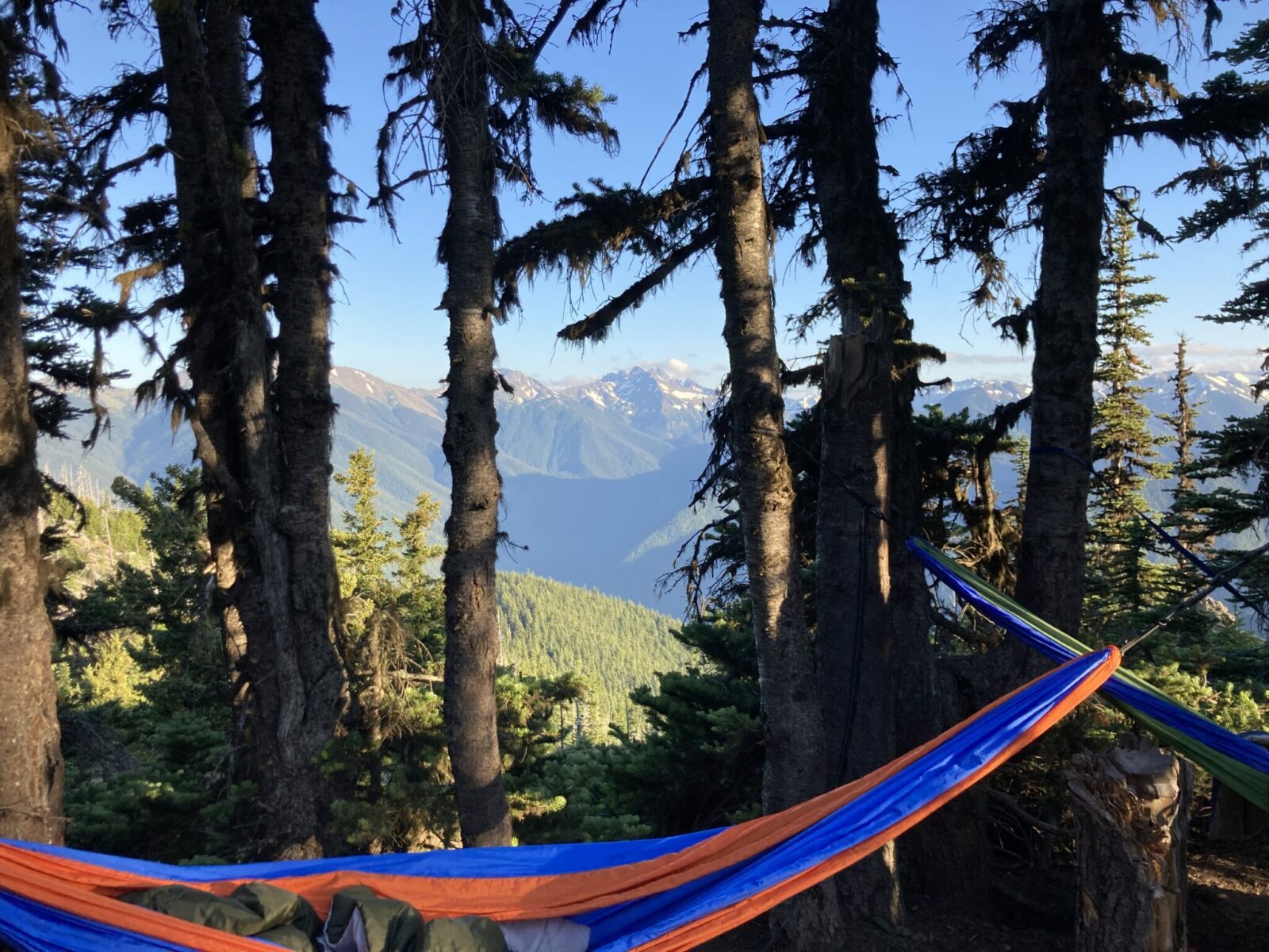 An orange and blue hammock and a green and blue hammock hanging from trees in the Deer Park campground. In the distance is a forested valley and high mountains with lingering snow