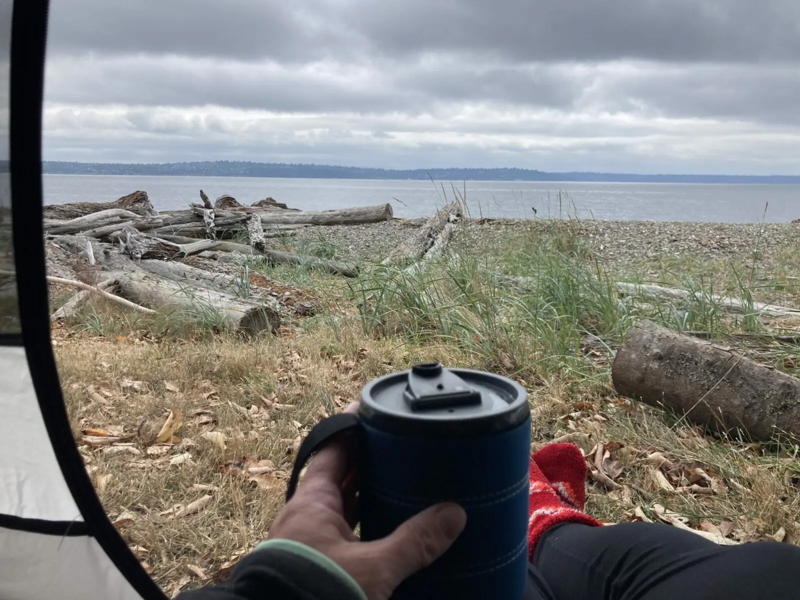 A person sitting in the opening of a tent at the beach. It's a cloudy morning and the person is holding a coffee cup and wearing leggings and red socks. There is distant forested land across the water