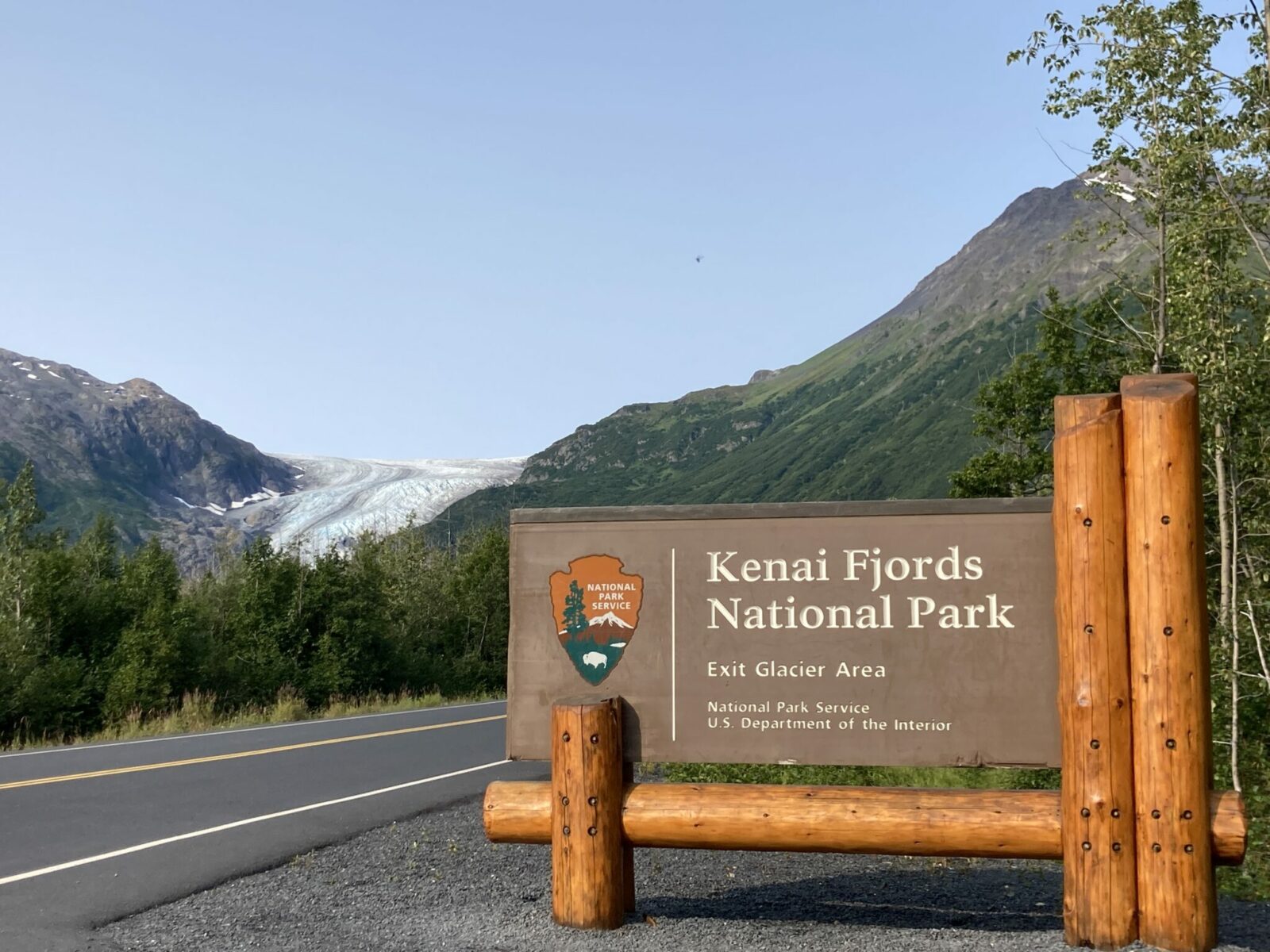 A sign saying "Kenai Fjords National Park Exit Glacier Area" on a brown national park service sign held up by logs. The sign is next to a two lane road going through a valley between green mountains towards a glacier which you can see in the distance.