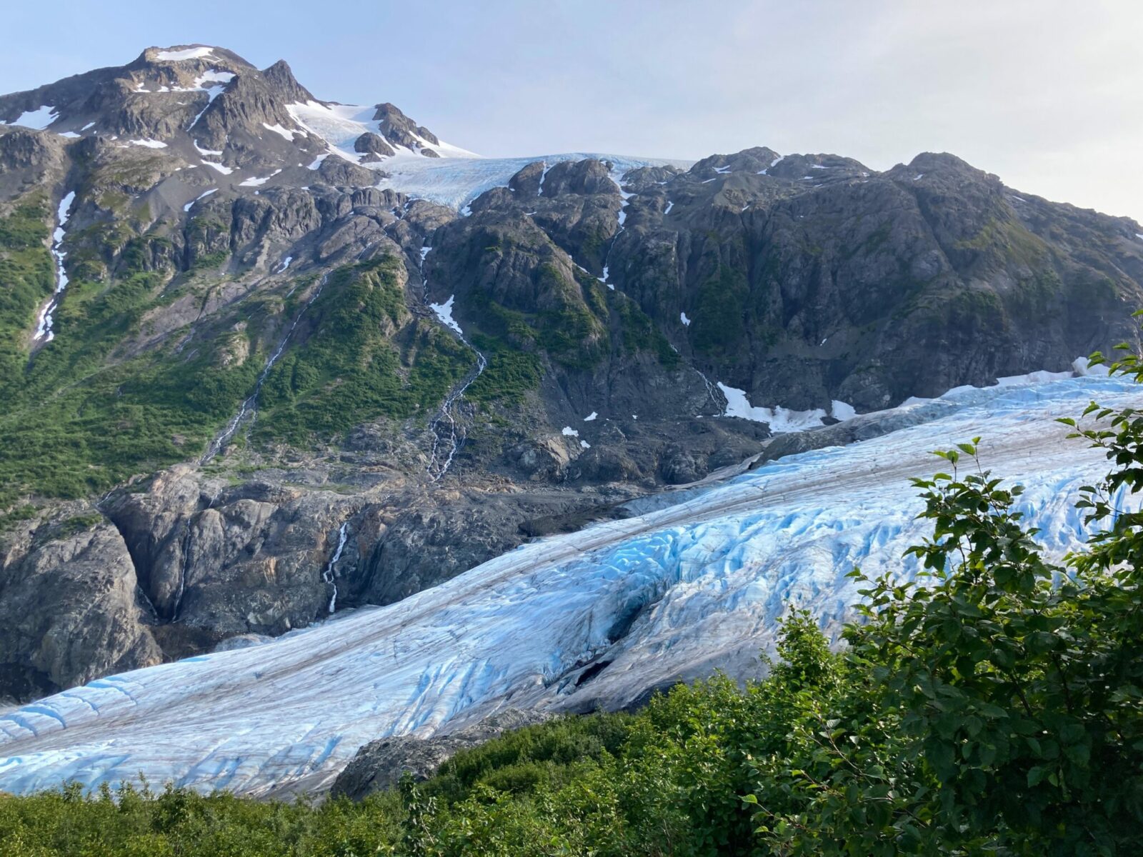 A glacier going between green brush and rocky mountains. The glacier is bright blue with dark brown streaks along the top